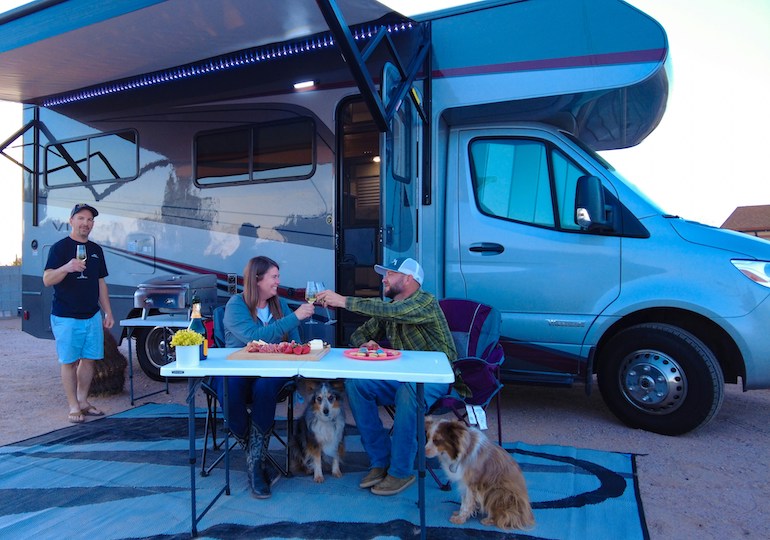 Dogs sat under a table where owners are enjoying a drink in front of a motorhome